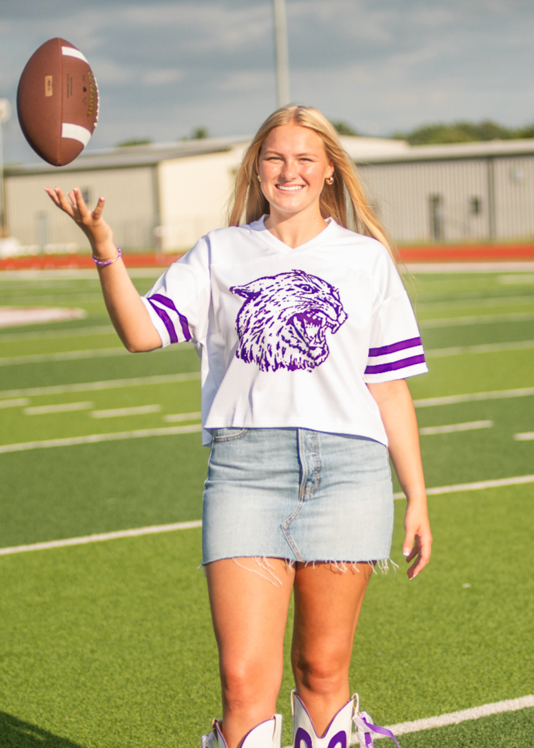 Person on a football field wearing a white shirt with a logo, denim shorts, and white boots with purple designs, holding a football.