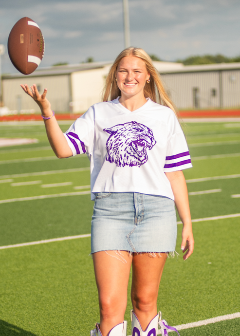 Person on a football field wearing a white shirt with a logo, denim shorts, and white boots with purple designs, holding a football.