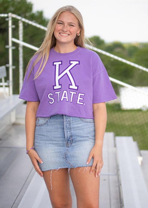 Person wearing a purple 'K State' t-shirt standing outdoors on a bridge.